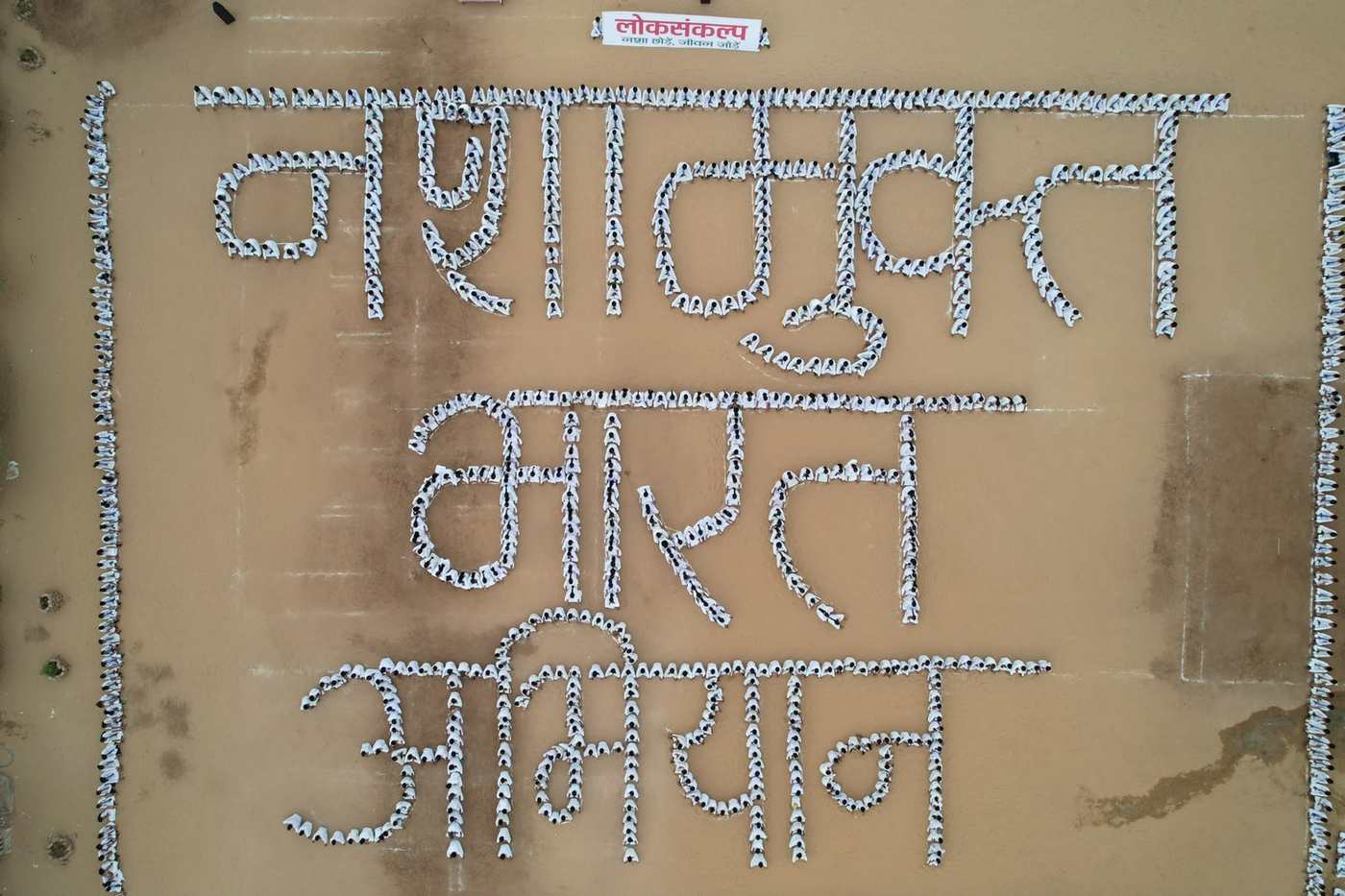 विद्यालय के मैदान में सैकड़ों विद्यार्थी सफ़ेद वस्त्रों में कतारबद्ध होकर “नशामुक्त भारत अभियान” के अक्षरों की आकृति बनाते हुए, ऊपर से लिया गया दृश्य