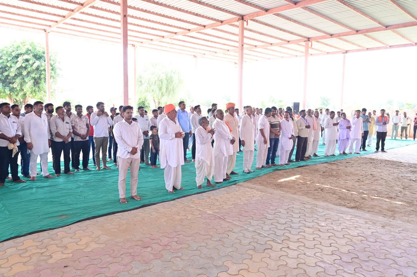 शेड के नीचे हरी दरी पर खड़े ग्रामवासी