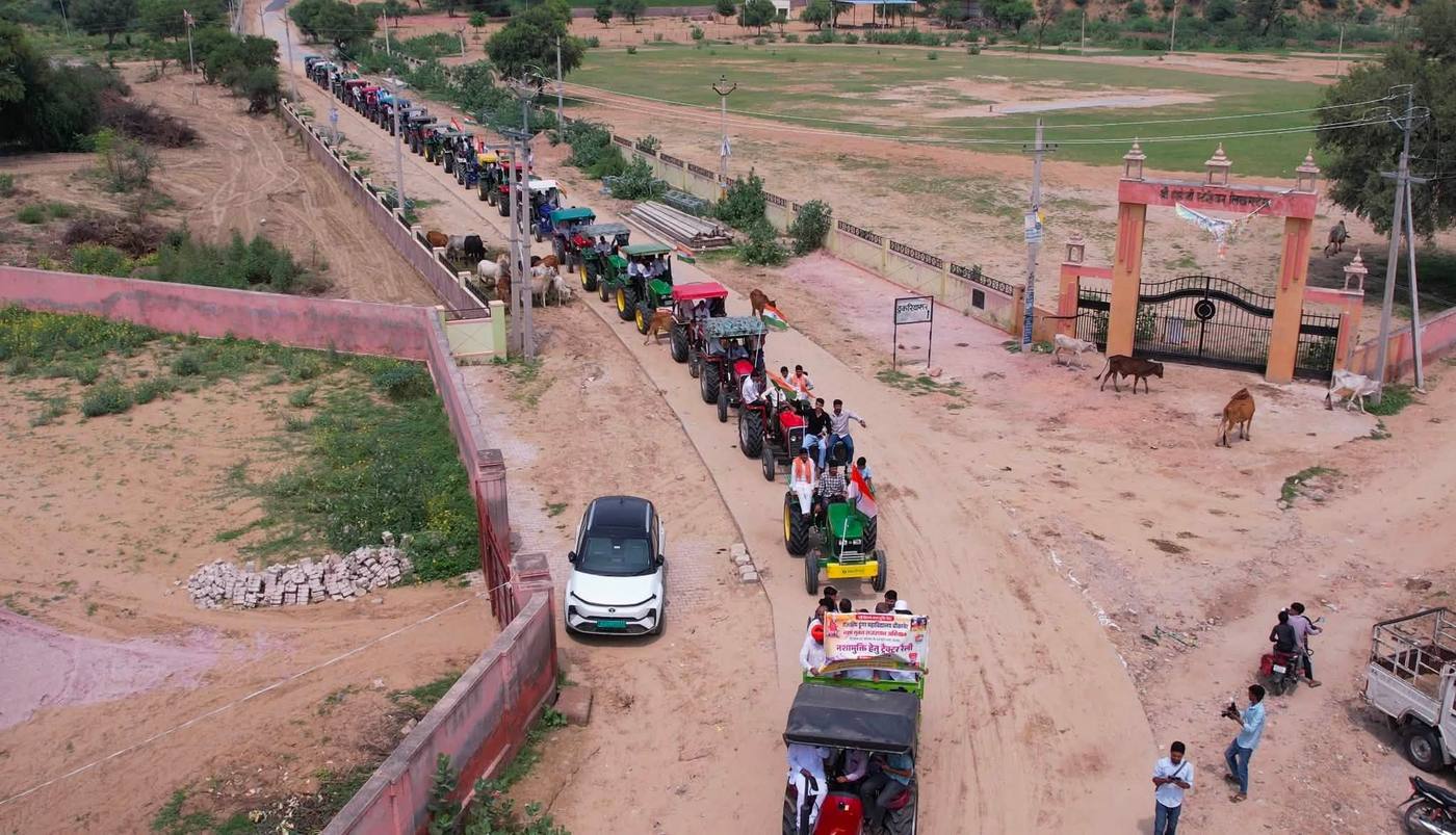 गाँव की सड़क पर कतार में चलते ट्रैक्टर, आगे नशामुक्ति रैली का बैनर, ऊपर से लिया गया दृश्य